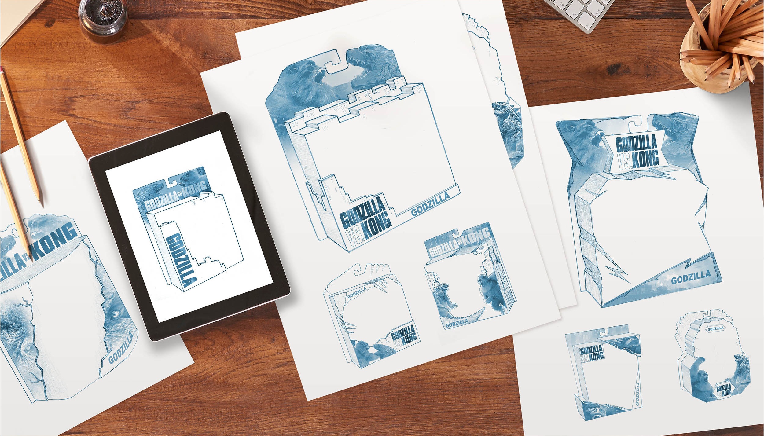 Several blue pencil sketches of Godzilla vs Kong toy packaging concepts are spread out on a wooden desk. The sketches show different box shapes and layouts, each labeled with Godzilla vs Kong and Godzilla. An electronic tablet displays one of the sketches. The scene feels creative and energetic, suggesting a design brainstorming session.